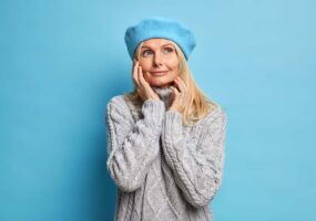 Portrait of pleased dreamy woman touches face gently looks away with peaceful expression wears beret grey knitted sweater isolated over blue background thinks of pleasant memories isolated on blue